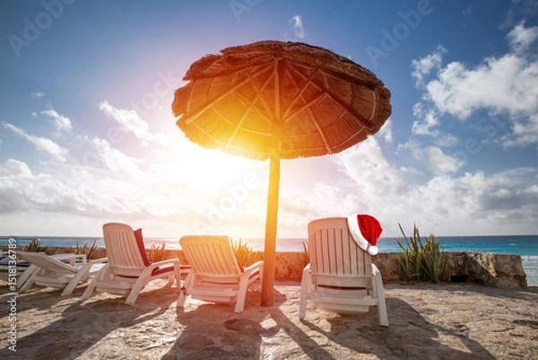 Obraz Santa Claus hat on sunbed on caribbean beach with grass umbrellas and chaise-longues. Christmas celebration