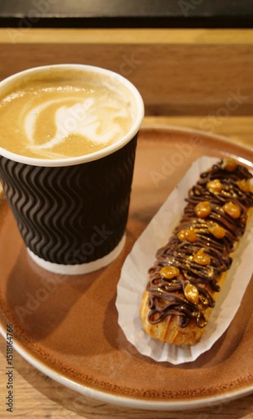 Obraz Classic Choco Eclair Sprinkled with Nuts and Coffee Latte in Paper Cup on a Clay Plate on a Wood Table