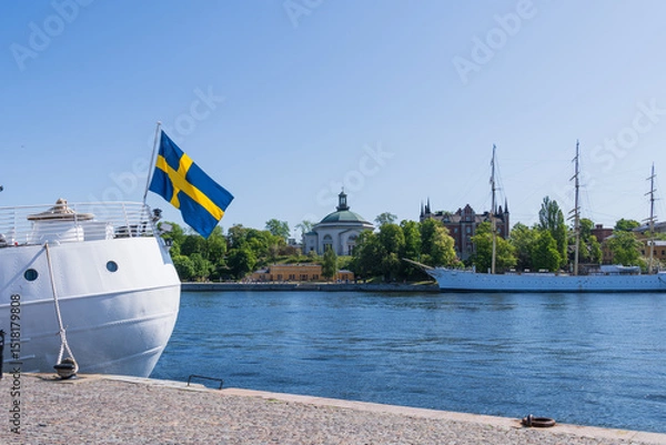 Fototapeta Stockholm, Sweden, swedish flag infront of the island of Skeppsholmen, with historical buildings and sail boats.
