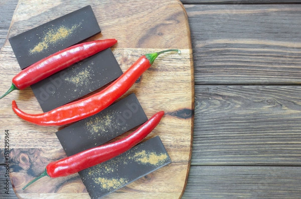 Fototapeta Bar of extra dark chocolate with three red chili peppers on a wooden cutting board on the table. Trend food concept. Selective focus. Horizontal orientation. Copy space. Top view