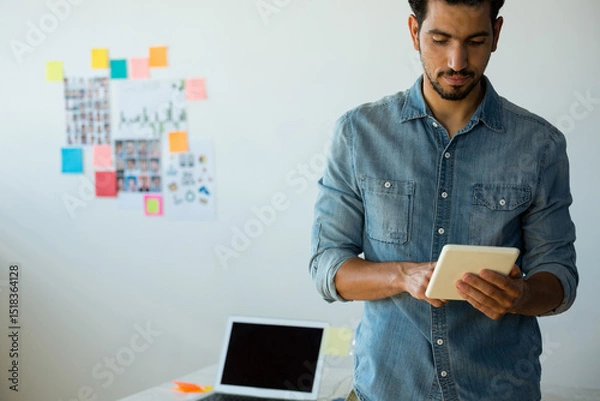 Fototapeta Middle Eastern pro standing reviewing tablet in modern office with sticky notes collage and laptop