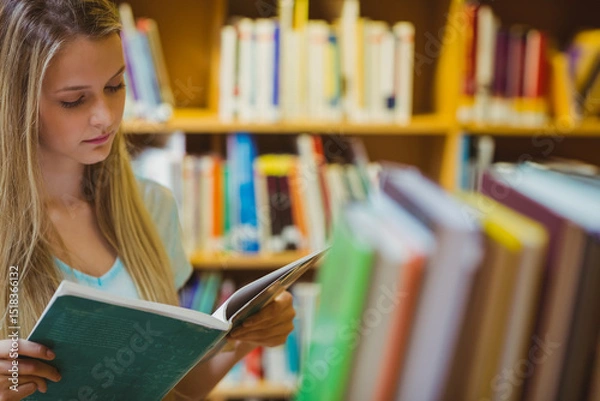 Fototapeta Teenage girl reading open book in library aisle with bookshelves and blurred books, copy space