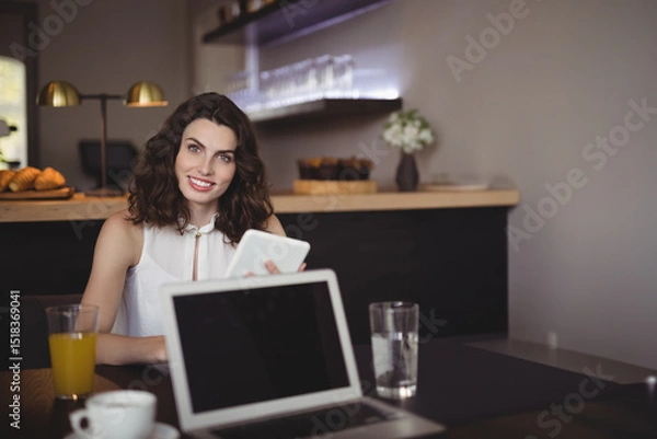 Fototapeta Woman in early thirties holding tablet, smiling at coffee shop table with laptop and orange juice