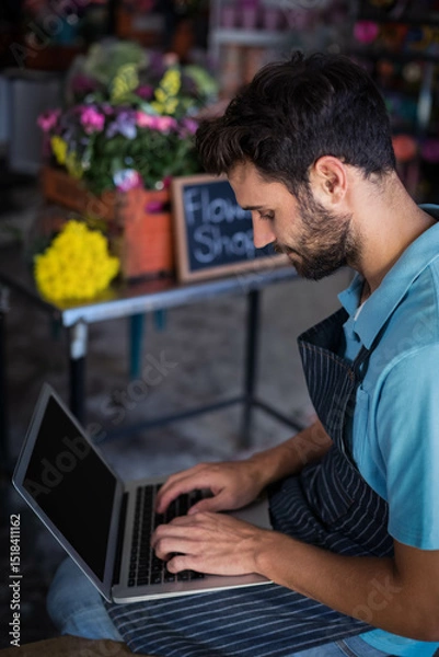 Obraz Bearded florist typing on laptop at flower shop workspace with wooden crates of colorful blooms