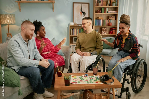 Fototapeta Diverse group of young adults and middle aged men and women with disability sitting together around board game table celebrating birthday, smiling and interacting in living room
