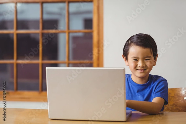 Fototapeta Asian boy sitting at wooden table and chair using laptop near multi-pane window smiling at camera