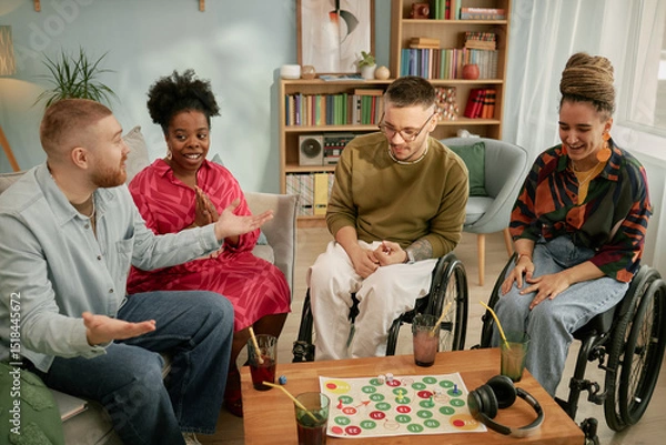 Fototapeta Diverse group of young adults including Black woman and Caucasian man and woman with disability in wheelchairs sitting together around table playing board game, during birthday celebration