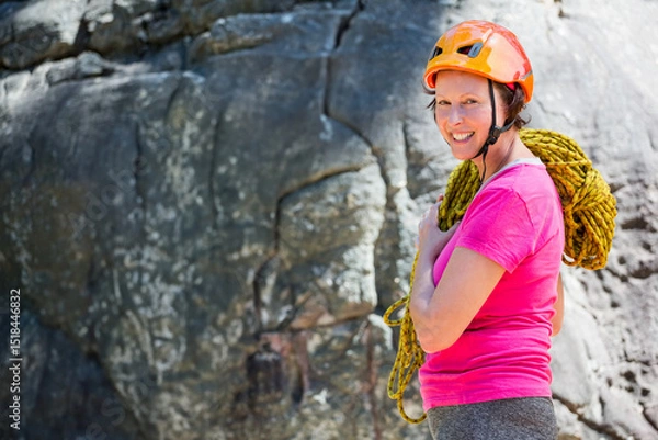 Fototapeta Woman standing at base of climbing cliff wearing orange helmet and carrying yellow rope, copy space