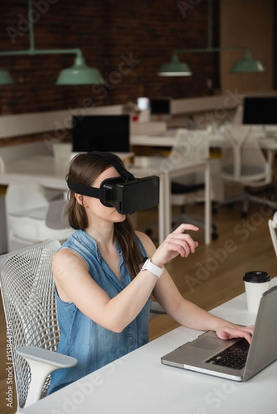 Obraz Female employee is wearing VR headset and sitting at white desk, with laptop in open-plan office