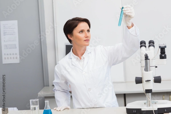 Fototapeta Female scientist in lab coat examining blue liquid in test tube at bench with glassware, microscope