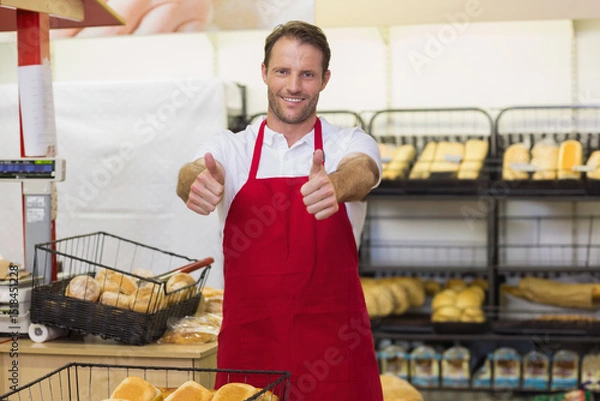Obraz Middle-aged baker wearing red apron smiling and giving thumbs up in store bakery section with bread