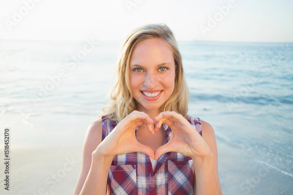 Obraz Woman smiling, making heart shape with hands at beach shoreline wearing button-up shirt