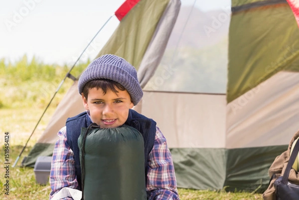 Fototapeta Middle Eastern boy sitting on grass at campsite holding sleeping bag near khaki-red canvas tent