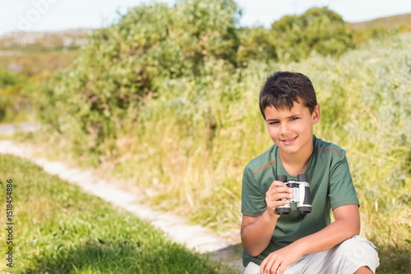 Fototapeta School-aged boy crouching beside dirt path in green shirt and shorts holding binoculars, copy space