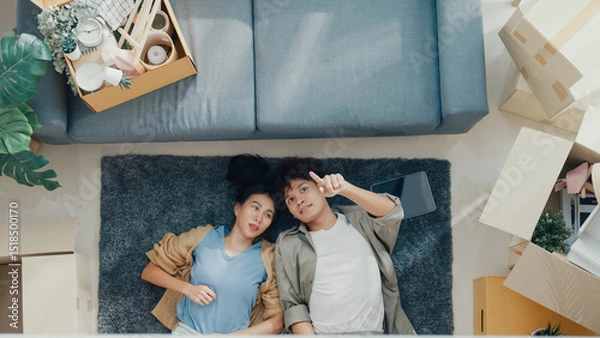 Fototapeta Top view. Happy Asian couple lay down and relax on carpet share idea for decoration furniture in empty area on living room at new house. Husband wife move house, Apartment Rent And Ownership Concept.