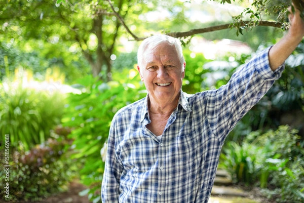 Fototapeta Senior man leaning on tree branch while smiling in backyard garden with stepping-stone path