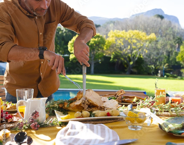 Fototapeta Middle-aged man carving roast turkey with knife and fork on patio table by pool, copy space