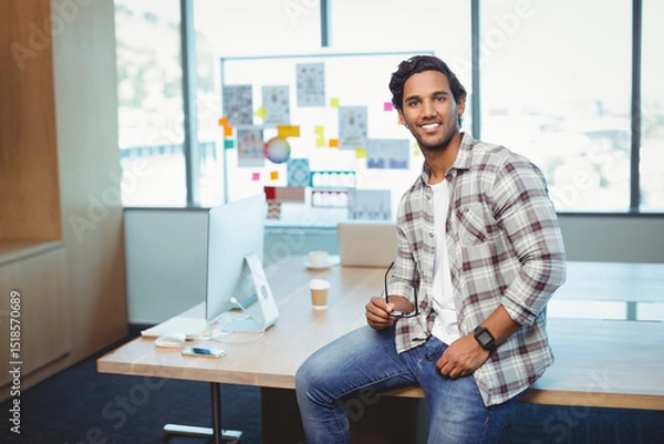 Obraz Indian man holding eyeglasses and smiling while sitting on office desk with laptop and coffee cup