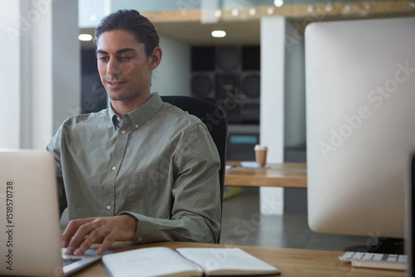 Fototapeta Middle Eastern man typing on laptop with notebook, coffee cup and speakers in office, copy space