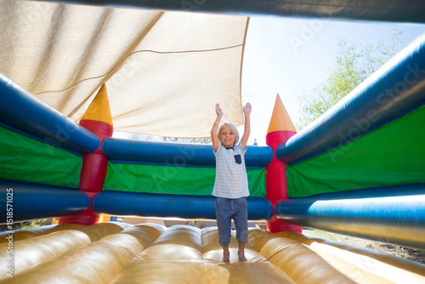 Obraz Boy bouncing inside brightly colored inflatable bounce castle under beige shade canopy in backyard