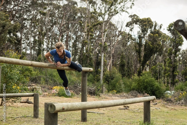 Fototapeta Woman vaulting over wooden beam in clearing on obstacle course wearing blue top, black leggings