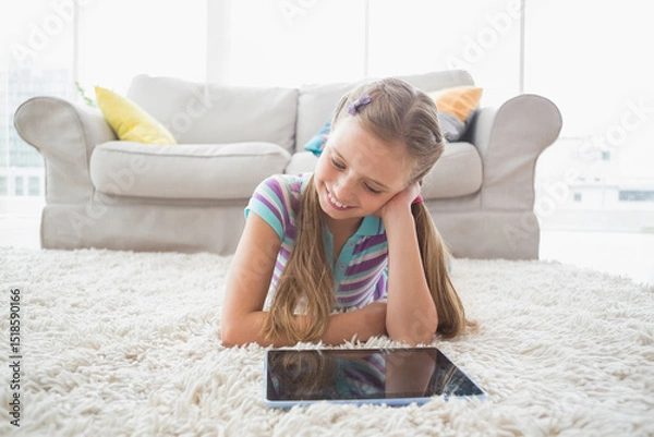 Fototapeta Girl lying on white shag carpet in front of gray sofa with colorful pillows using tablet