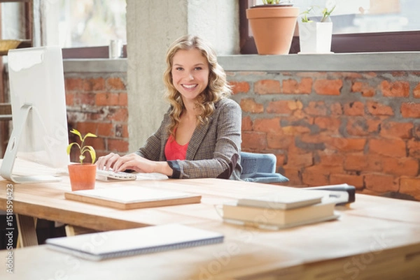 Fototapeta Teenage girl smiling while working at loft-style office desk with large monitor and potted plant