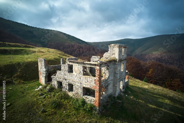Fototapeta Abandoned stone building on grassy hillside, partially in ruins. Structure against backdrop of rolling hills and dense forest, under sky with dramatic clouds, evoking sense of history and solitude.