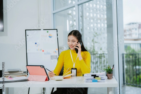 Obraz Image of young Asian woman, office worker busy with documents and smiling while holding a phone, office background.