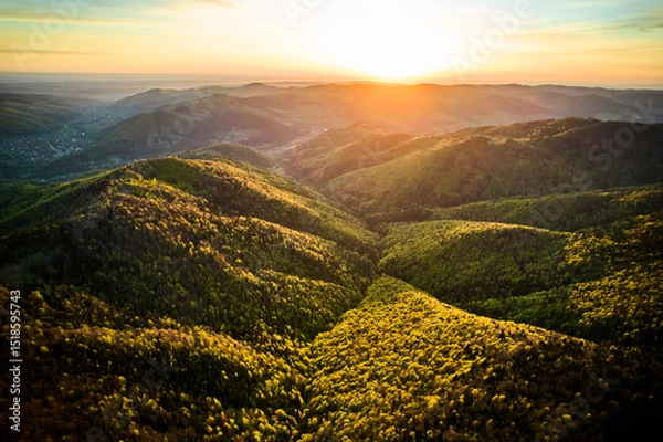 Fototapeta Aerial view of breathtaking sunrise over rolling mountains covered in mix of lush green and autumnal foliage, with dramatic clouds streaking across sky, casting warm, golden light over the landscape.