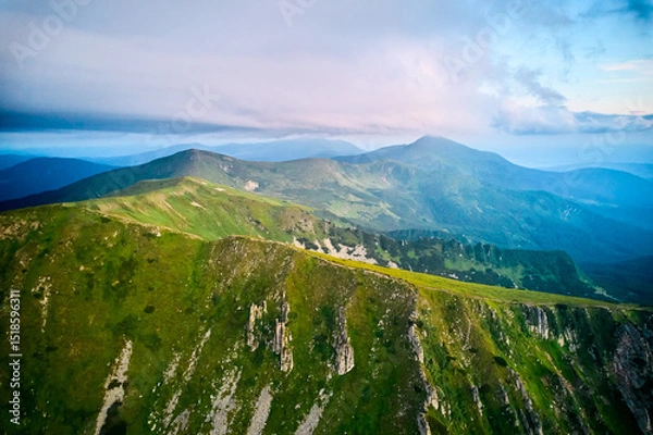Fototapeta Breathtaking aerial view of green, rugged mountain peaks under dramatic sky. Scene captures contrast between vibrant landscape and moody clouds, with sunlight breaking through. Carpathians, Chornogora