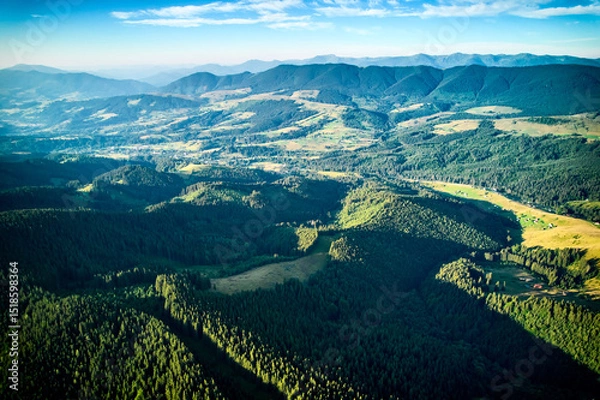 Fototapeta Aerial view of densely forested mountains stretching into distance under clear blue sky with scattered clouds. Landscape reveals valleys and distant fields, showcasing natural beauty of region.