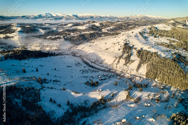 Fototapeta Aerial view of snow-covered valley with winding road passing through small village. Landscape features gently rolling hills, dense forests, and distant mountains under clear blue sky, with soft mist.