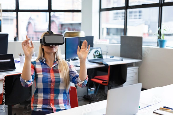 Fototapeta Office worker wearing VR headset raising hands at open plan office desk with laptop and monitors