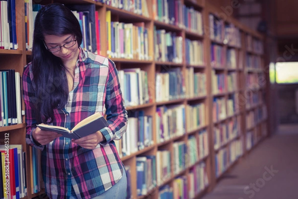 Obraz Student leaning against tall wooden bookshelf in library aisle, reading open book with glasses
