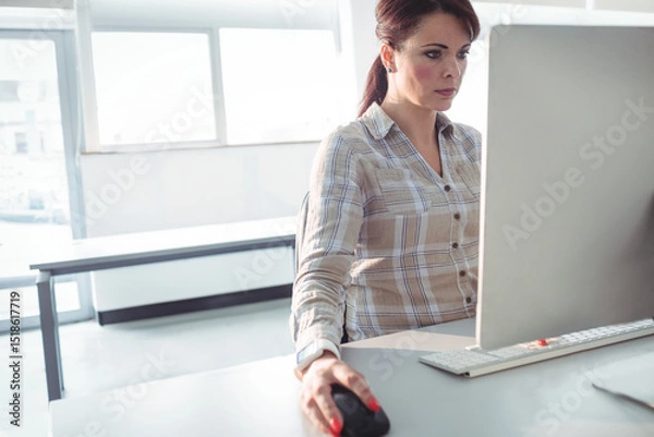 Obraz Woman wearing plaid shirt sitting at office desk using computer near windows, bench, copy space