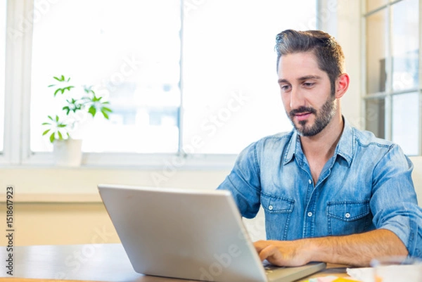Obraz Man typing on silver laptop at wooden desk in bright office with potted plant, copy space