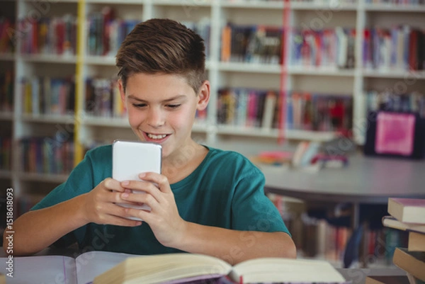 Fototapeta School-age boy sitting at library table holding smartphone reading open book with stacked books