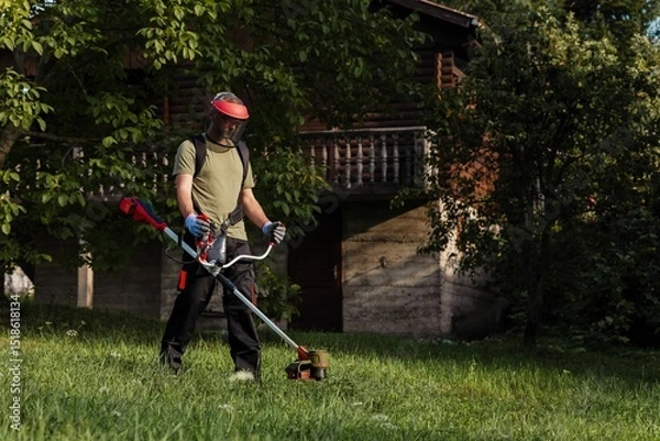 Obraz Man using a trimmer to maintain grass near a wooden house during a sunny day