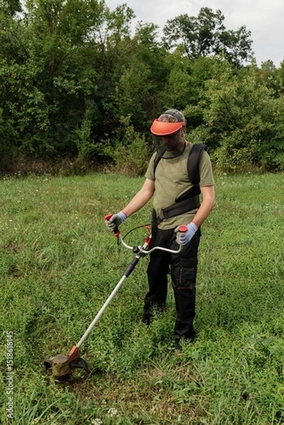 Obraz Man using a string trimmer in a grassy field surrounded by trees on a sunny afternoon