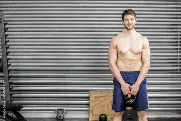 Obraz Man gripping kettlebell at thigh level in gym with weight rack, plyometric box, copy space
