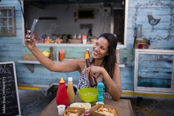 Obraz Woman taking selfie at wooden table in front of food truck holding beer bottle and smartphone