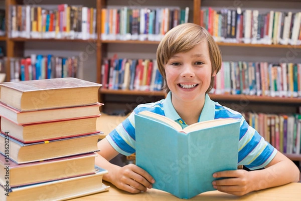 Fototapeta Preteen student reading light blue book at wooden library table, with stacked volumes behind