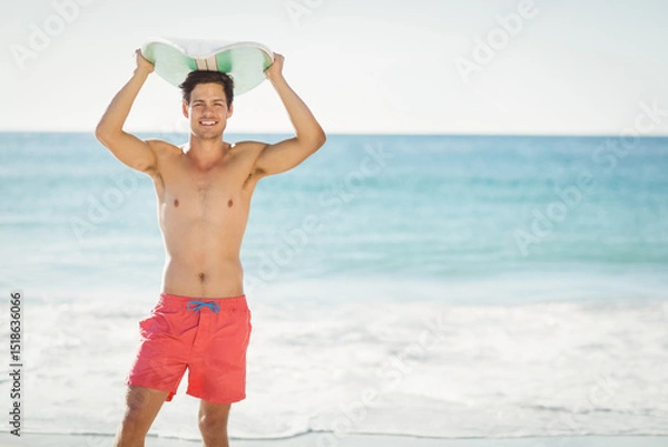 Obraz man standing barefoot on sandy beach holding surfboard overhead wearing red swim trunks