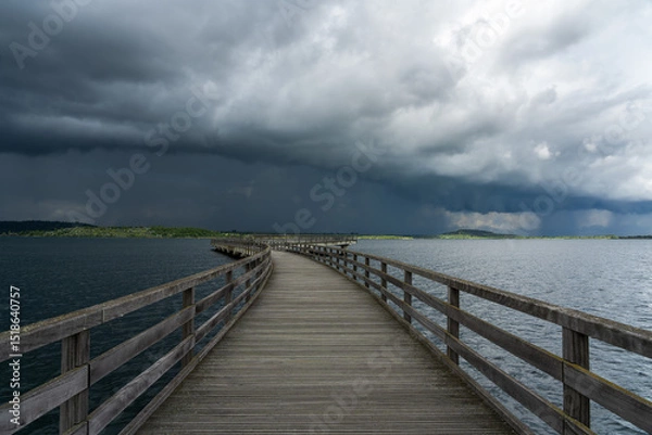 Fototapeta The photo shows a pier leading into calm waters, while dramatic storm clouds create a gloomy mood.