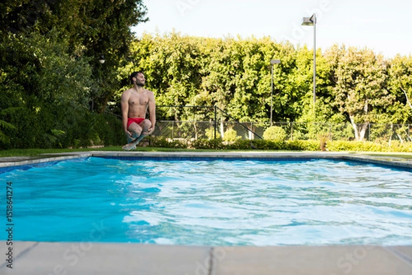 Obraz Fit man in red trunks crouching at concrete pool edge preparing to dive into blue water