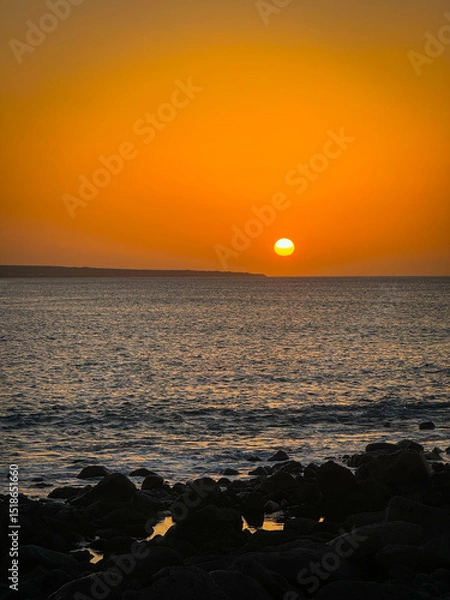 Fototapeta Brilliant orange sun dips towards horizon, casting a warm glow across the sky and reflecting on gently rippling ocean waves. Dark volcanic rocks create a silhouette against serene and fiery sunset.
