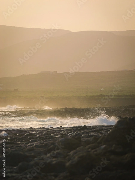 Fototapeta Waves crash against dark, rocky volcanic shore and disappear into a misty, ethereal haze that envelops distant hills. Muted tones and soft lighting create an atmospheric, mysterious coastal landscape.