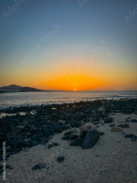 Fototapeta Evening coastal landscape with rippling Atlantic Ocean and distant volcanic mountains. Setting sun bathes sky in warm orange and soft blue hues, illuminating vast stretch of rocky and sandy coastline.