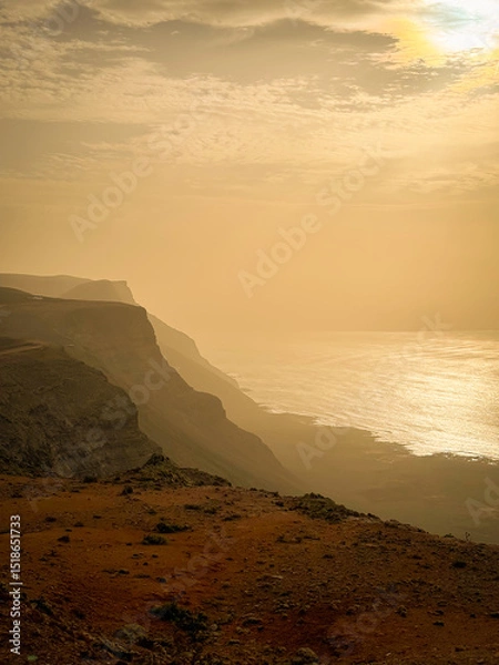 Fototapeta SILHOUETTE: Dramatic coastal cliff disappears into the sunlit, misty ocean. Shimmering water contrasts with dark, layered landforms to create powerful and atmospheric landscape under soft, cloudy sky.